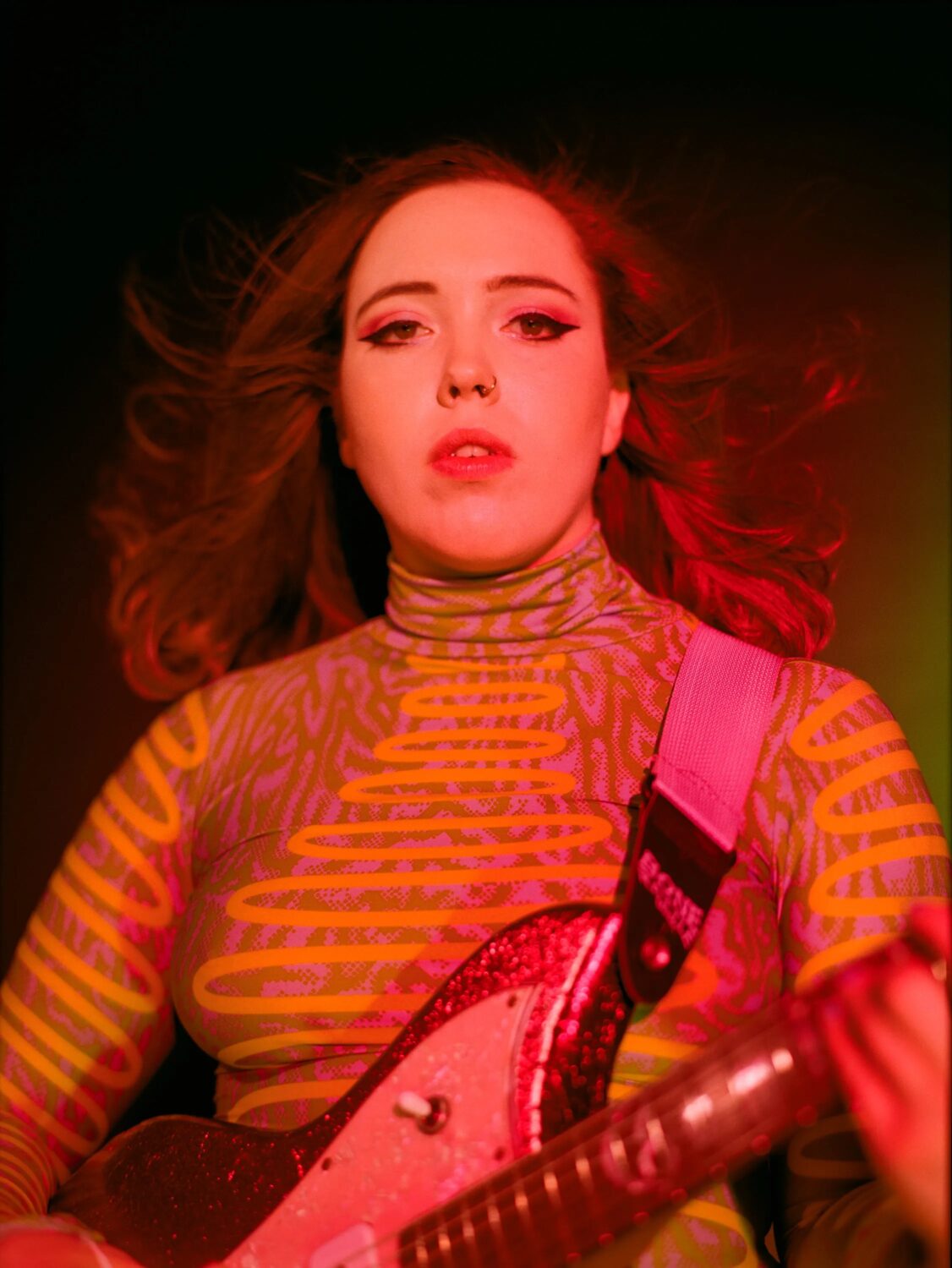 Woman wearing a geometric yellow patterned tight turtleneck shirt basks in red stage light and holds a guitar